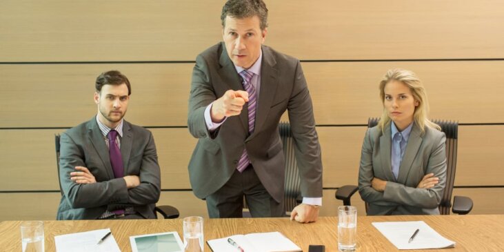 Three business people at a table with paper, one standing and shaking a finger at the person across from them