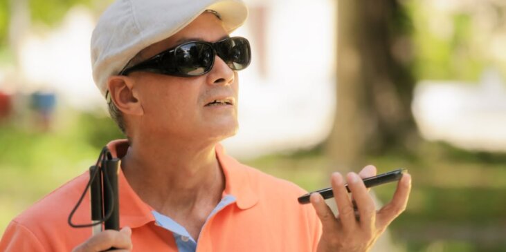 A man wearing sunglasses and a cap holds a smartphone and a white cane outdoors.